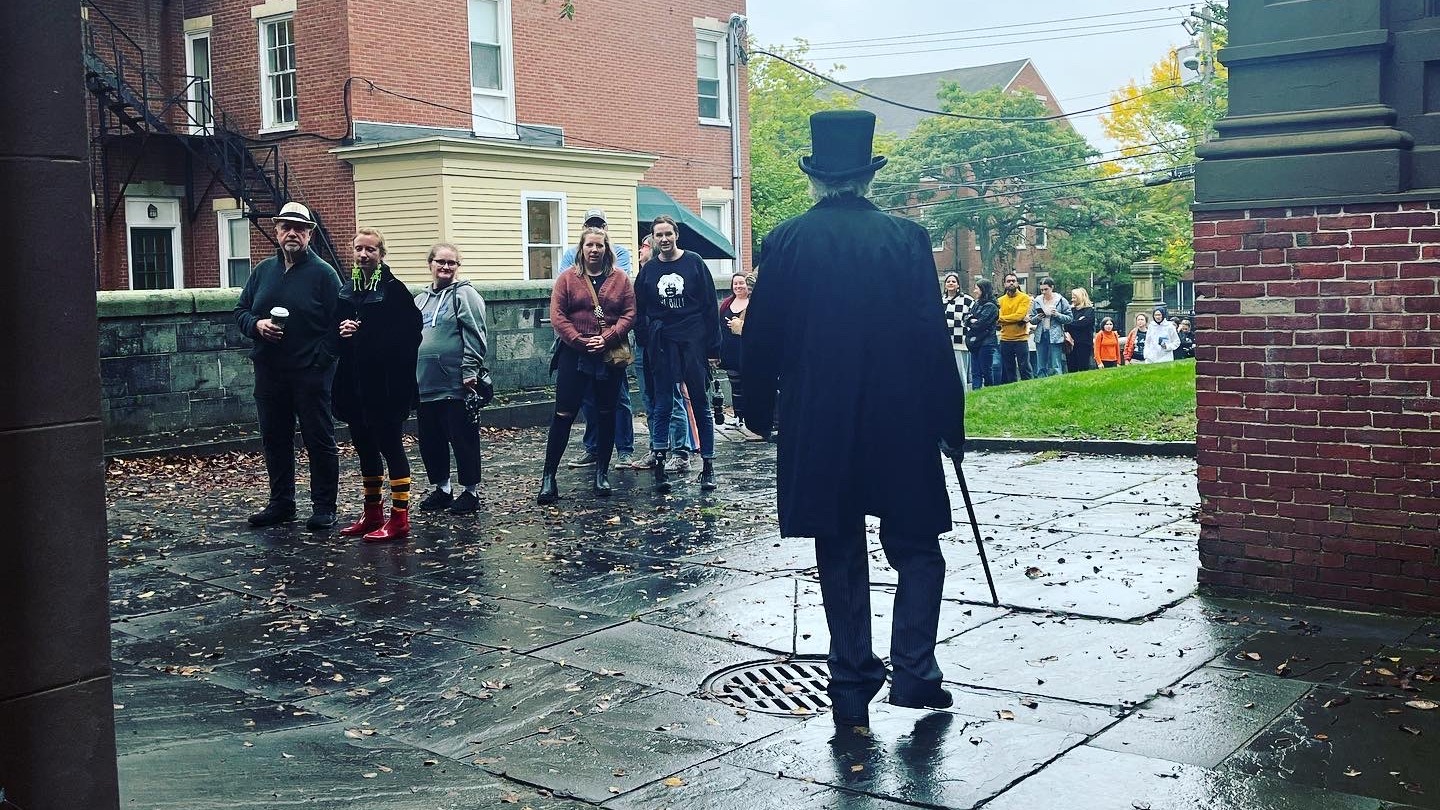 Actor Andrew Harris, dressed in a top hat and suit as Charles Dickens, walks into Victoria Mansion's cobblestone courtyard, where he is greeting a line of guests. The sky is grey and the weather is rainy.